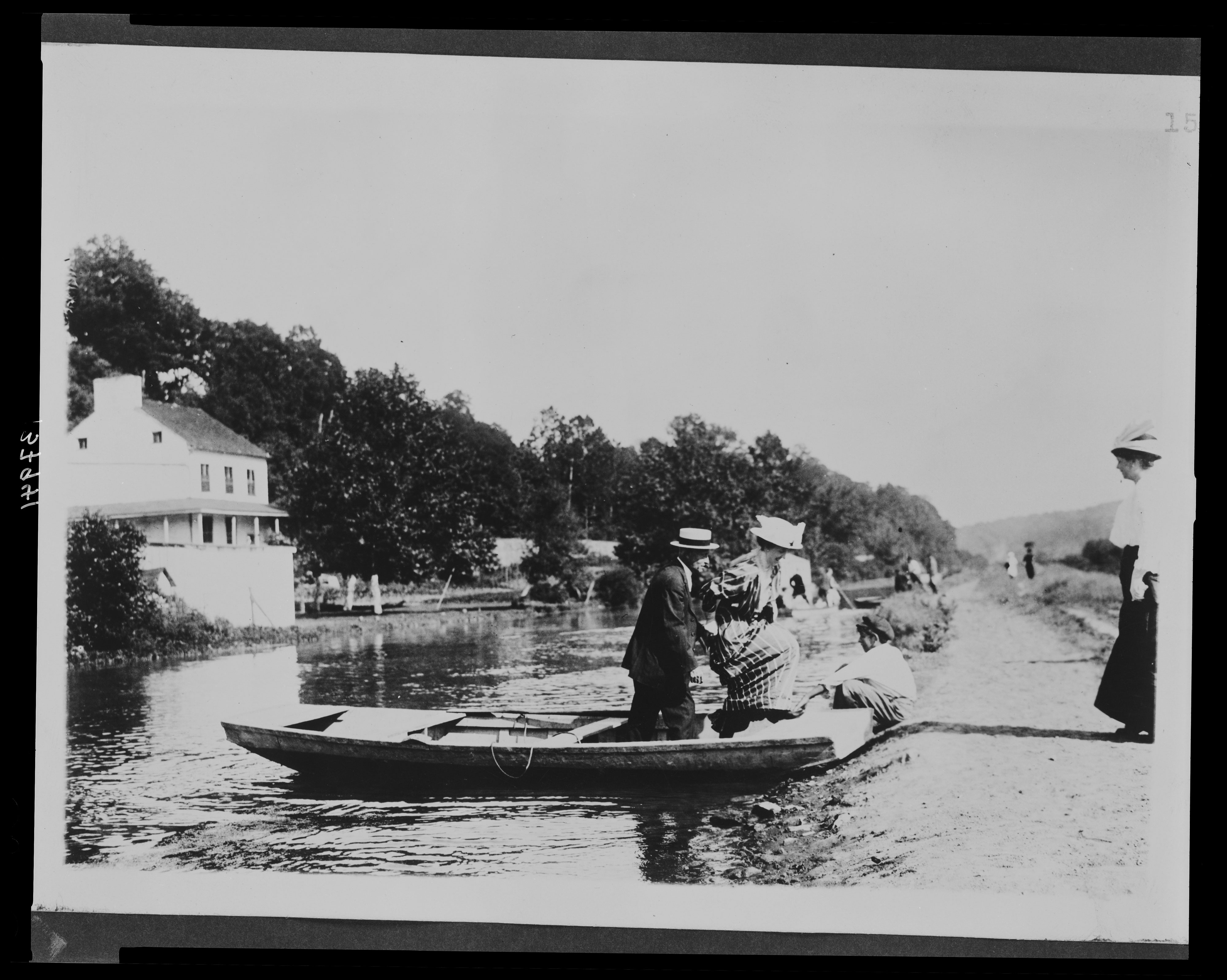 Black and white photo of two people stepping off a small boat on the Chesapeake and Ohio Canal. A young boy is holding the boat against the towpath. A woman stands to the right watching them. The Abner Cloud House can be seen on the left side of the image