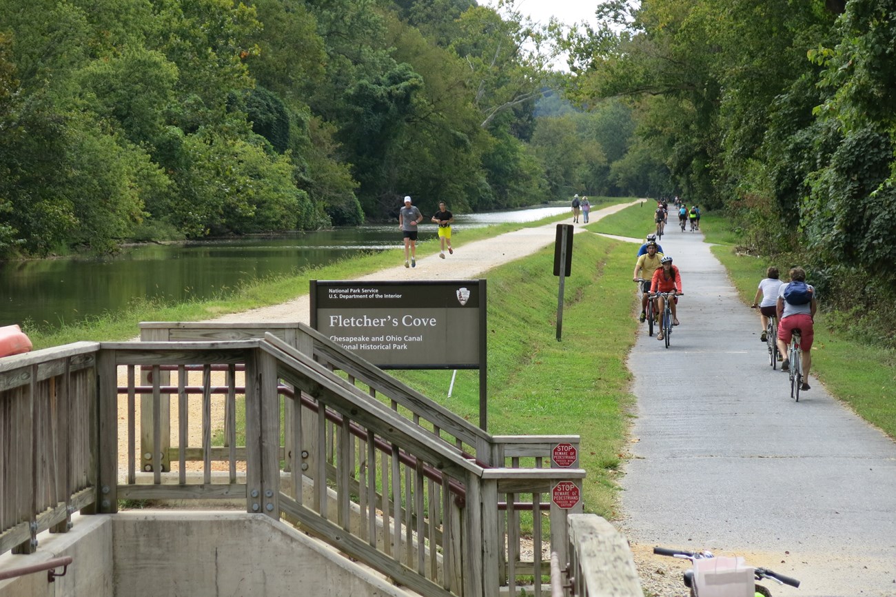 CHOH Capital Crescent Trail in 2015 Color photograph showing two parallel trails along the water-filled canal. People walking on a gravel trail is on the far left, while people riding bicycles on a paved path is at right. Steps associated with a wooden footbridge extend over the canal
