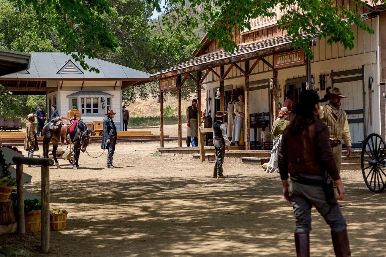 People in western period clothing in the dappled shade in front of a store front.