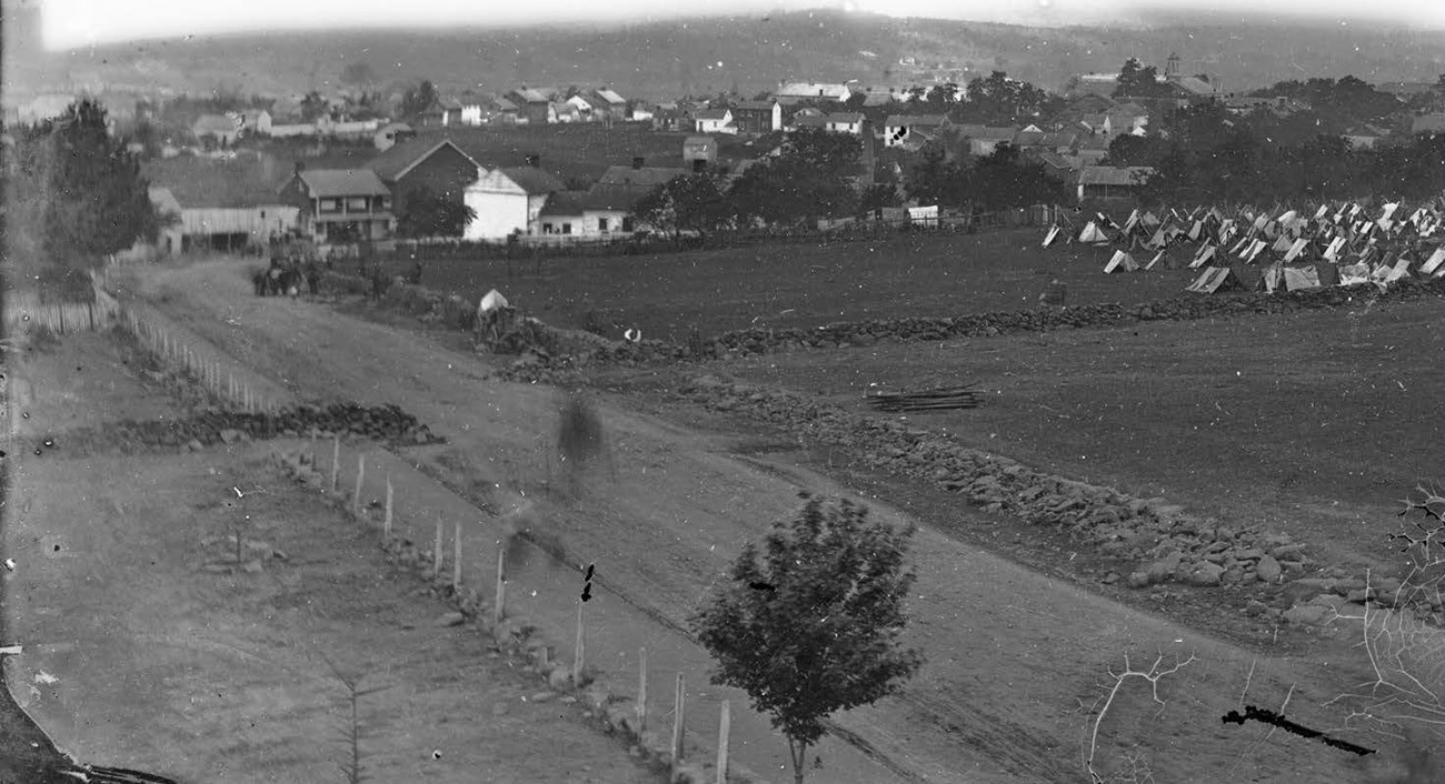 A high angle view of a low stone wall along a broad road, with a cluster of tents to the right.