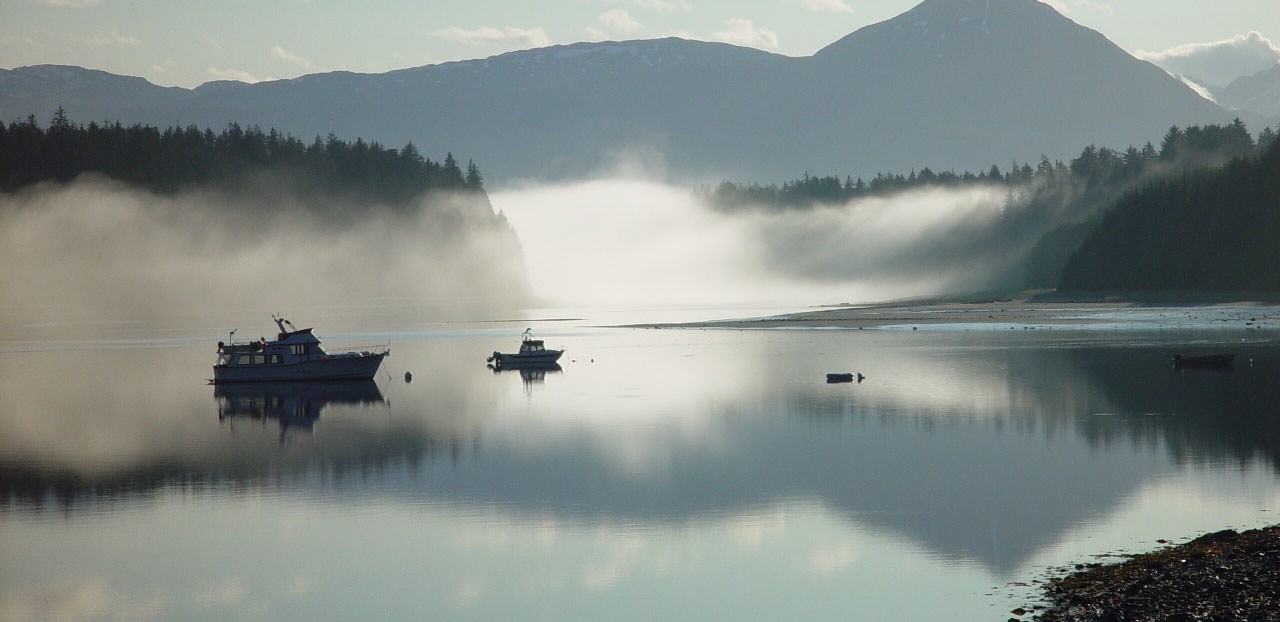 Low fog hangs in a cove, between mountains and still, reflective water where several boats float