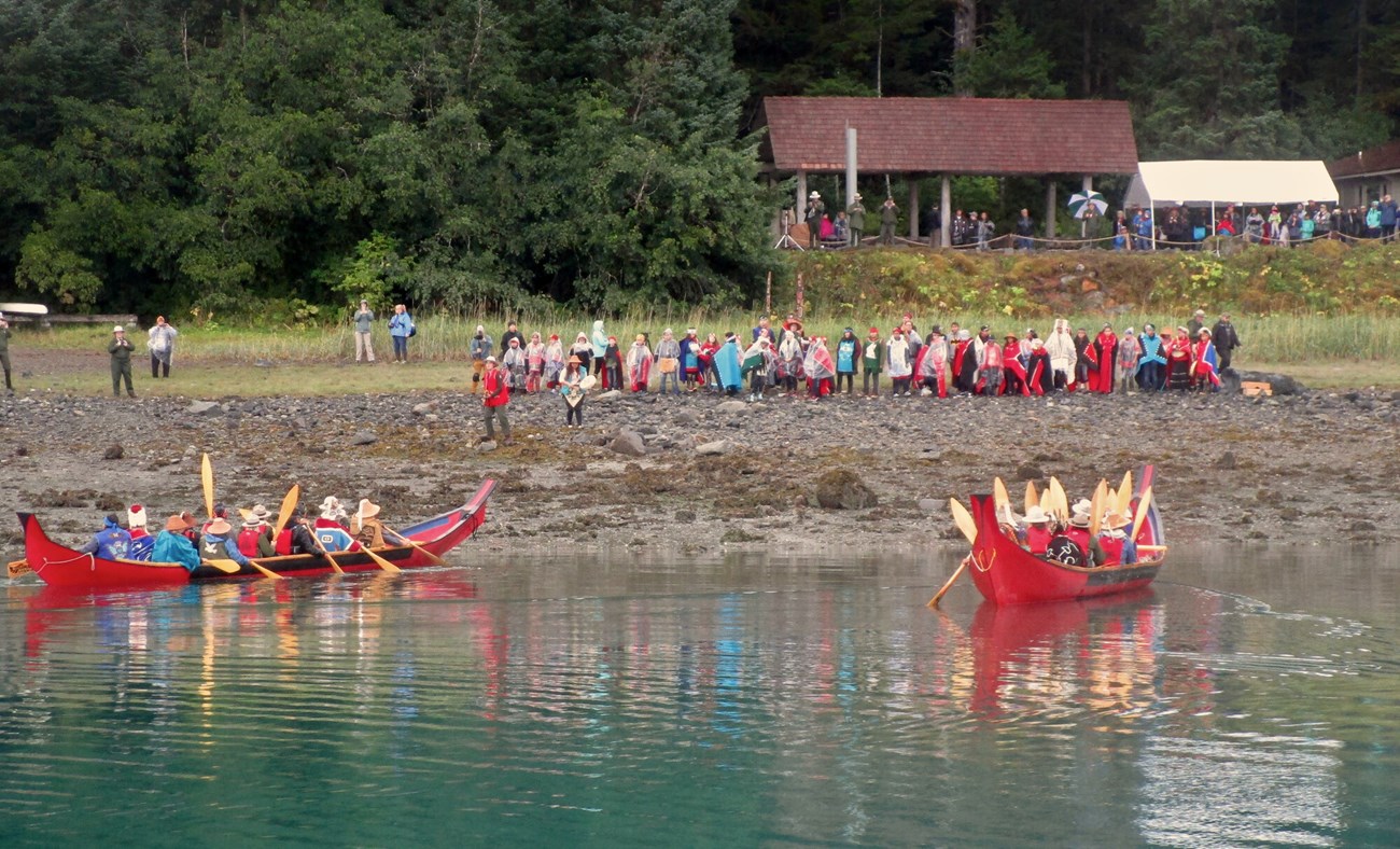 Bright red dugout canoes pull close to shore in Bartlett Cove while a crowd looks on and a Tlingit ceremony takes place.