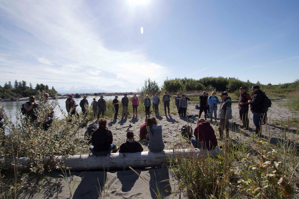 A group stands in a circle on a beach with low vegetation