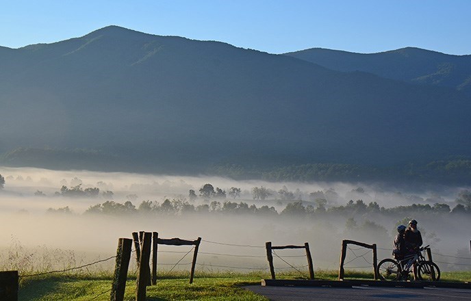 Two people with bikes beside a wood and wire fence, with a fog-filled valley and hills beyond