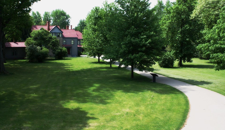 A row of leafy trees and turf grow alongside a walkway that leads towards a large house with chimneys.