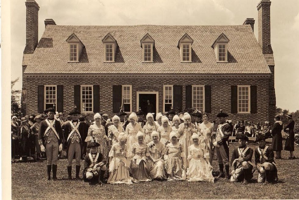 People in colonial costume stand in two rows in front of a brick house.