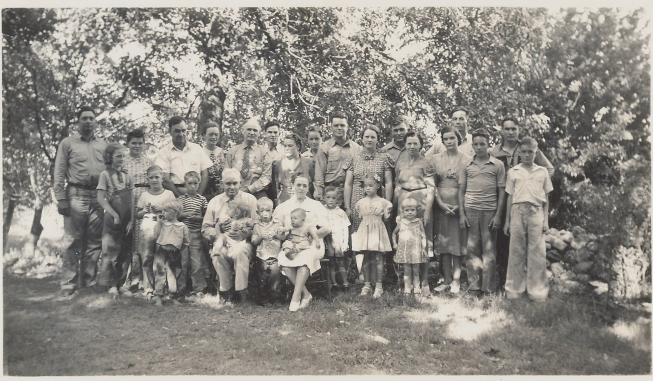 A group of people of various ages stands and sits in rows to pose for a photo, outdoors in front of orchard trees.