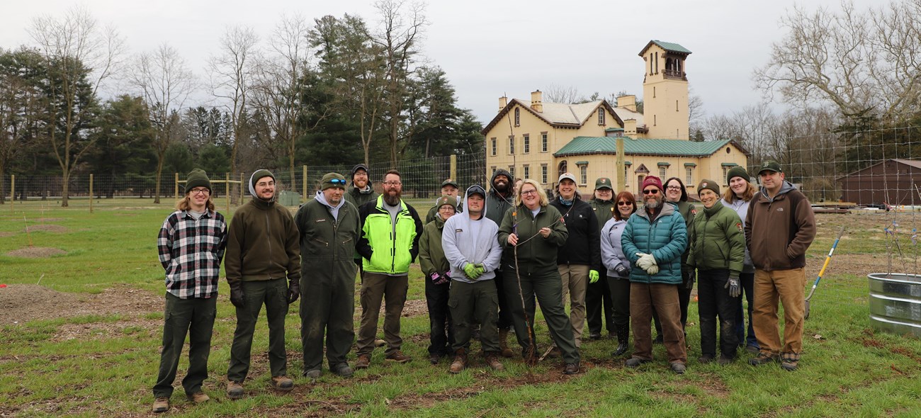 A group of 19 NPS staff, volunteers, and partners in hats, coats, and work gear pose in the orchard area in front of the mansion.