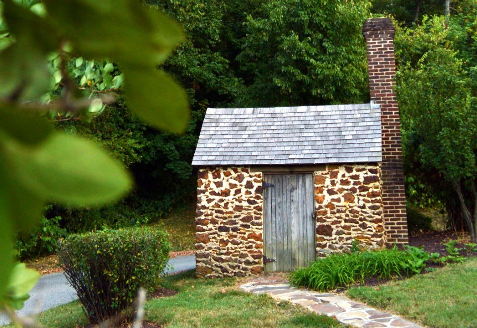 Lush green vegetation surrounds a small stone structure, with a wooden door and brick chimney