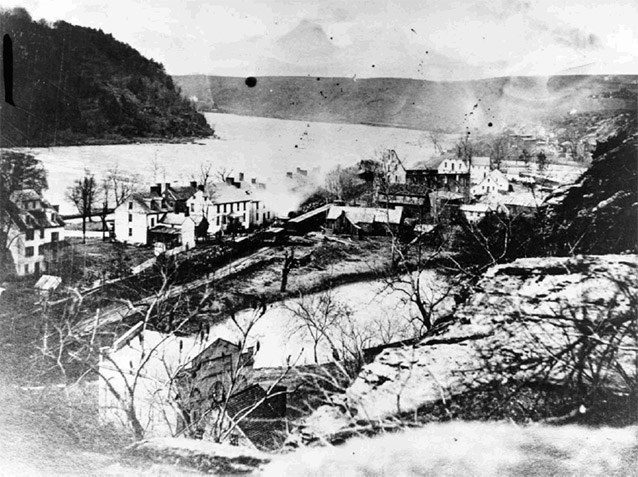 Black and white photo from a hillside shows residential and industrial structures on an island, with people on train cars passing through the middle.