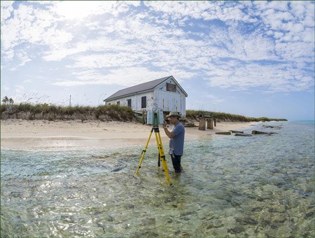 lt= A man stands in knee-deep water near a sandy beach, using a laser scanning device on a tripod.