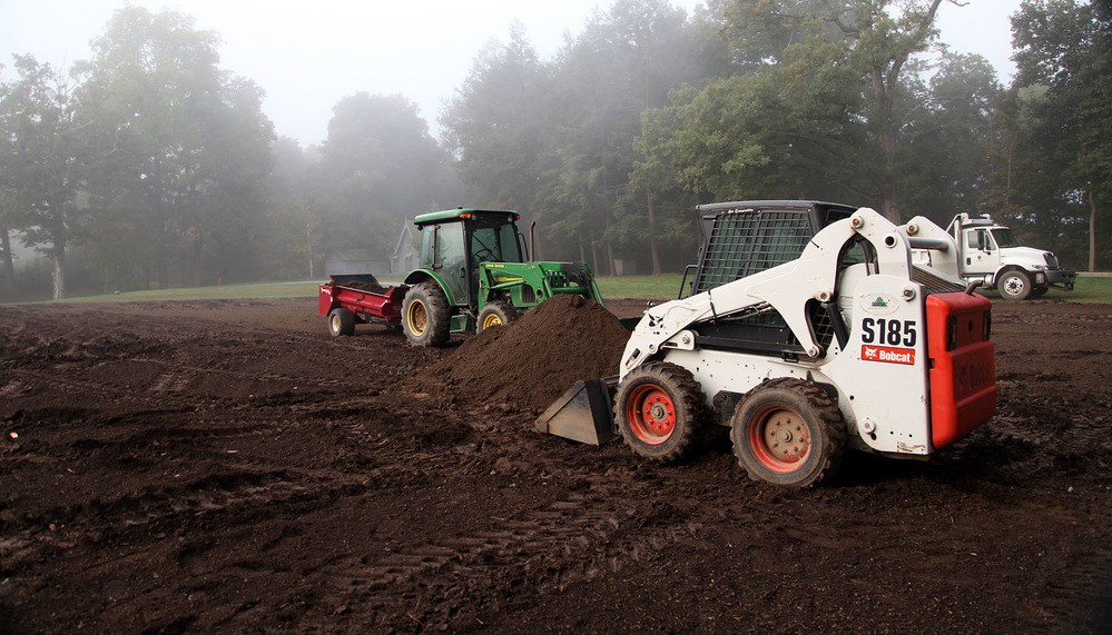 A heap of fine, dark topsoil stands in the middle of an expanse of dirt, with a backhoe and a tractor.