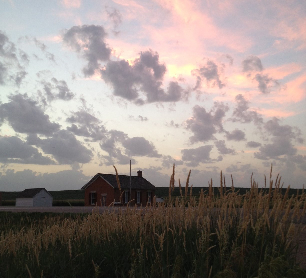Soft pink and blue sunset glowing behind puffy clouds over the one-room Freeman School, behind tall grasses