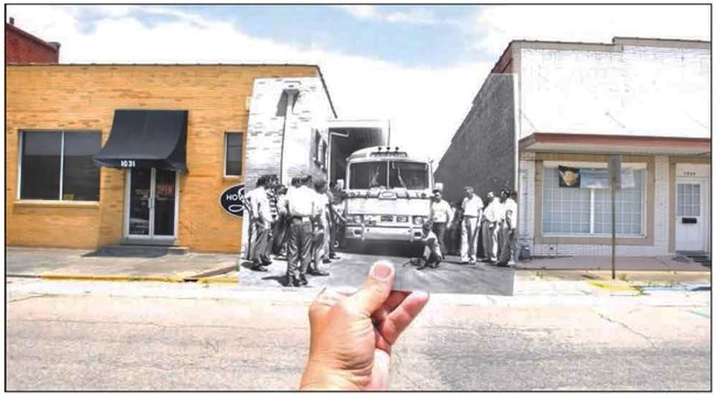 A 1961 photograph showing a group of people standing near a bus, in an alley between two one-story brick buildings.