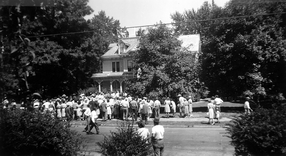 A crowd of people on the sidewalk and yard in front of a Victorian house.
