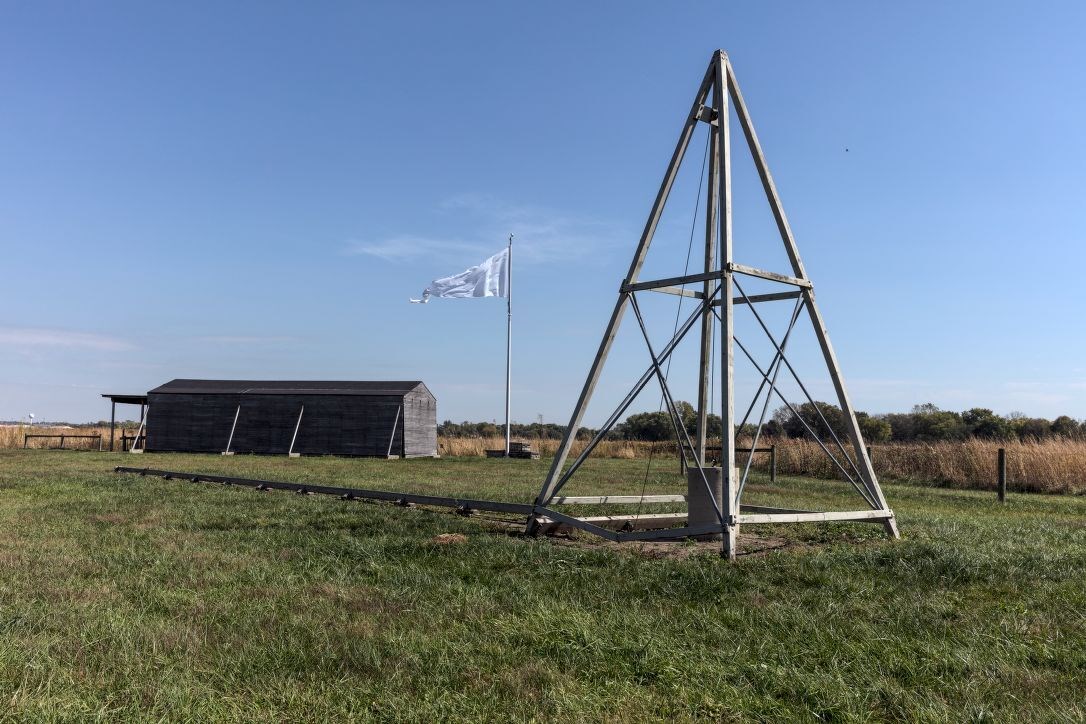 Pyramid shaped catapult in an open field, with a wooden hanger and white flag in the background
