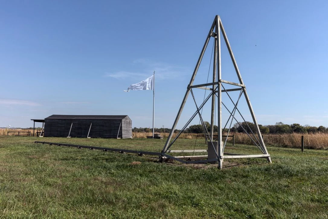 HUFFMAN FIELD CATAPULT II Pyramid shaped catapult in an open field, with a wooden hanger and white flag in the background