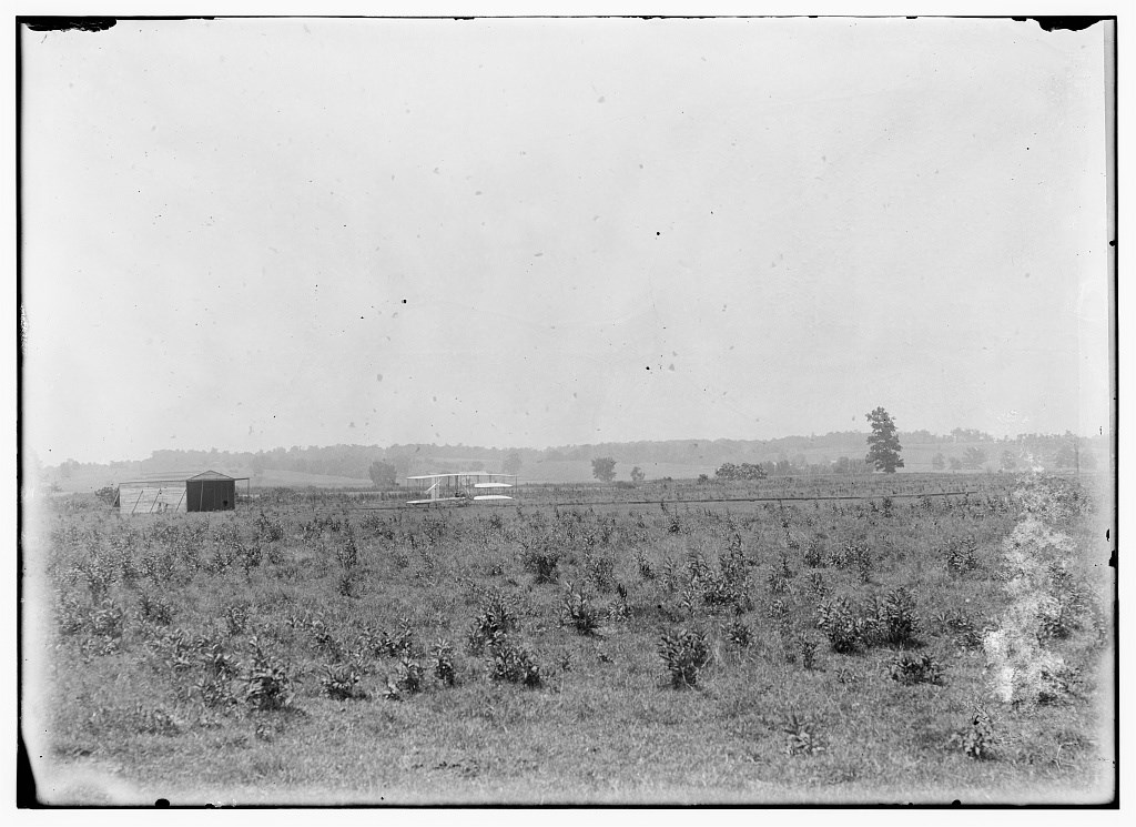 Long-range view of machine on launching track, showing hangar nearby and hummocky ground of former swamp at Huffman Prairie, Dayton, Ohio