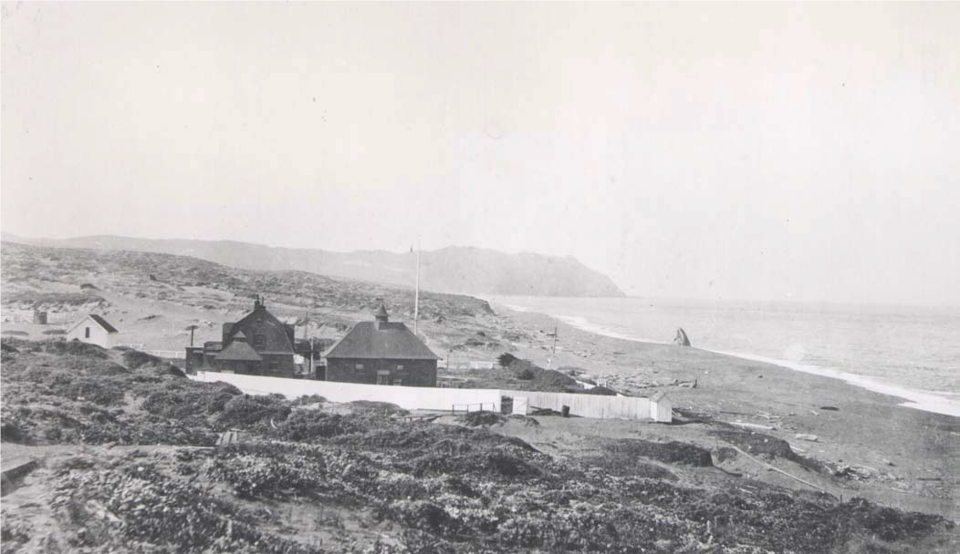 Black and white photograph of a few dark-sided buildings surrounded by a picket fence adjacent to an exposed ocean shoreline.