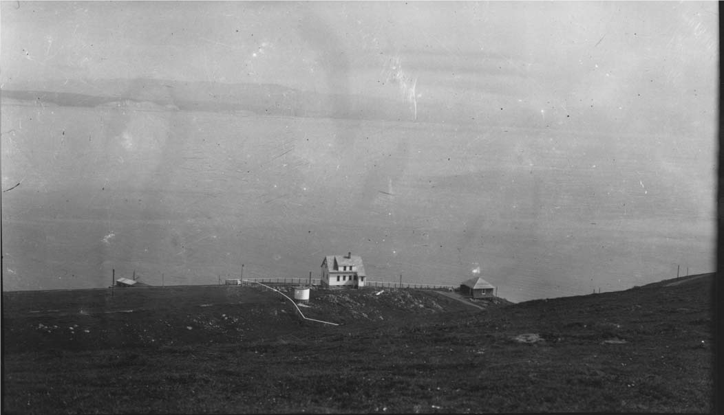 The Officer-in-Charge quarters on Drakes Bay, ca. 1928. A black and white photograph of a two-story house adjacent to a small two-car garage and a smallish white water tank, all clustered at the base of a grassy slope in the mid-distance above a bay.