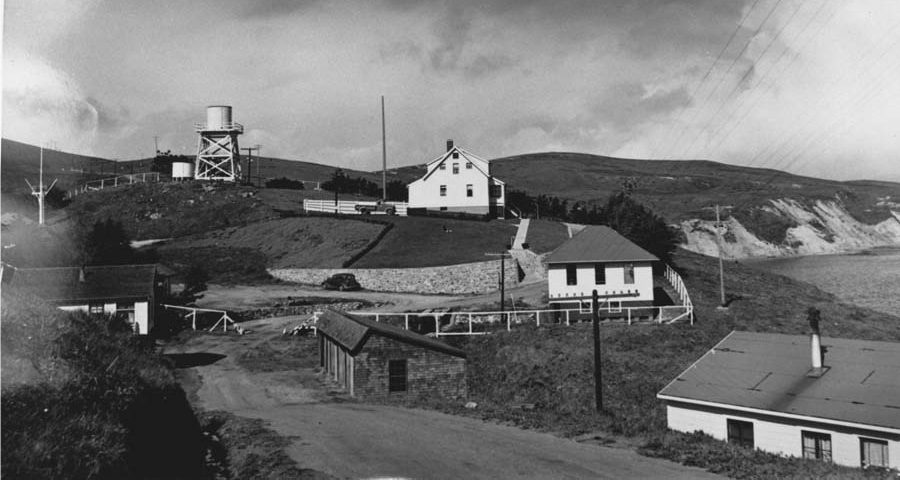 A black and white photograph of buildings built near the edge of a coastal bluff top. Above and beyond the buildings on the rolling hills to the left is a water tank tower. A narrow road passes between the buildings, partially along a rock retaining wall.