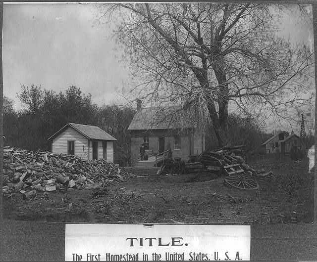 Photograph shows man standing on ramp in front of Daniel Freeman homestead in Gage County, Nebraska, the first homestead claim under the 1862 Homestead Act; large piles of wood in yard.