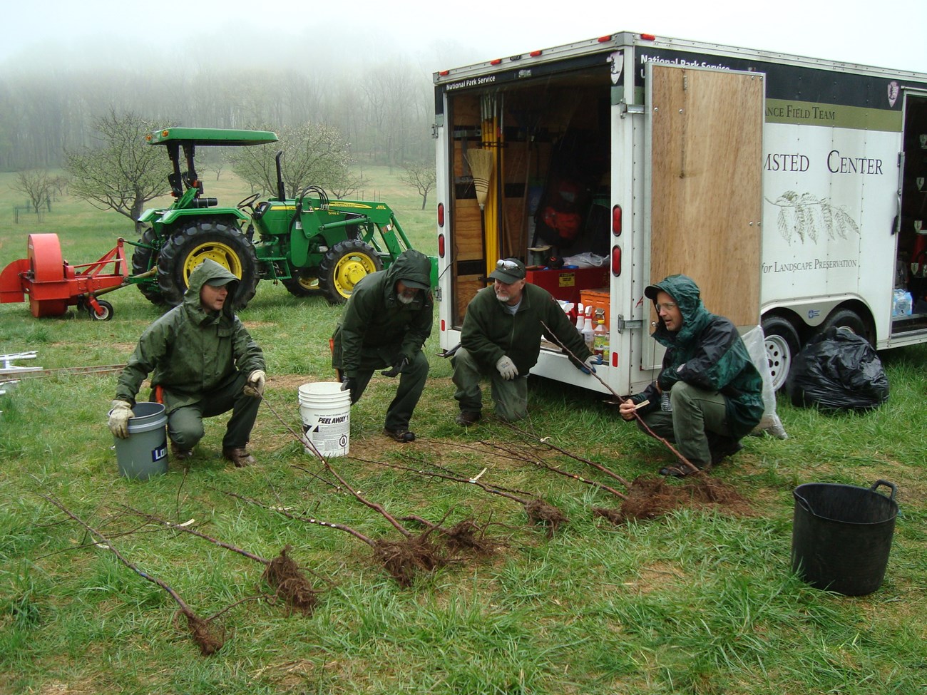 Historic Plant Nursery (OCLP) Four employees in raingear kneel beside a row of bare root trees on the ground behind a box truck.
