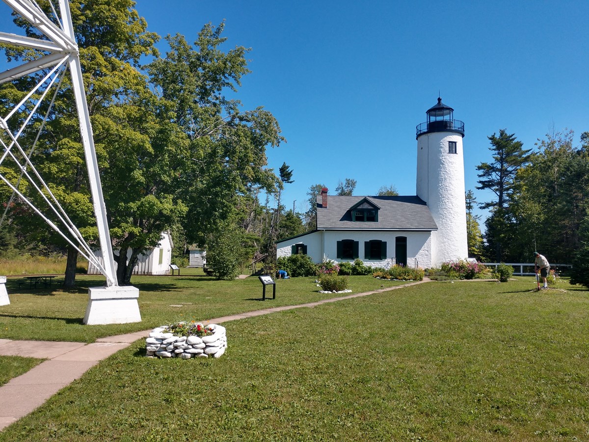 A tall, metal lighthouse tower stands in front of shorter brick lighthouse, connected by a walkway across turf and garden beds.