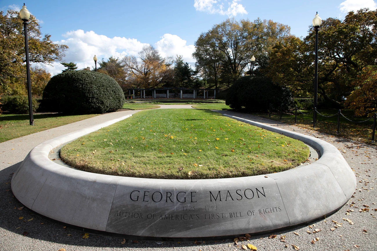George Mason Memorial Entrance The words “George Mason 1725-1792 Author of America’s First Bill of Rights” are engraved on the edge of a rounded concrete turf planter at the entrance of the George Mason Memorial. A sidewalk, manicured shrubs, and low trees surround the planter.