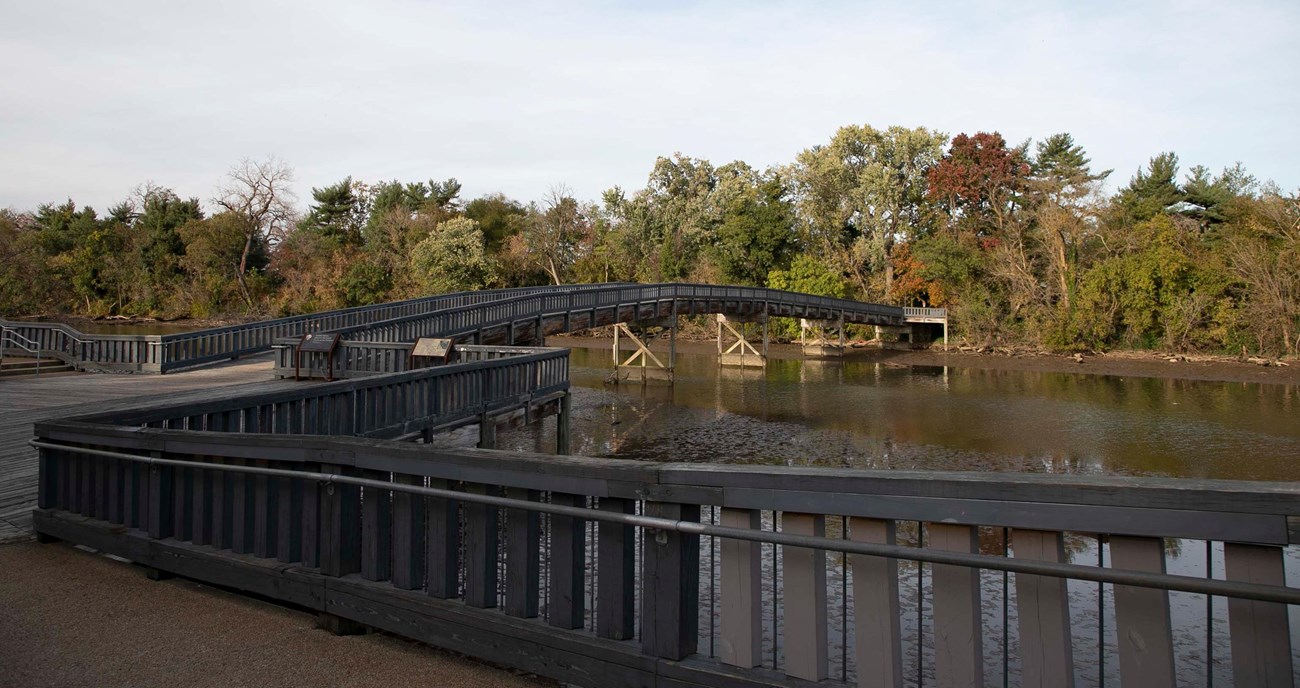 Pedestrian bridge over boundary channel A wooden pedestrian bridge arches slightly to cross a waterway towards a tree-lined shore.