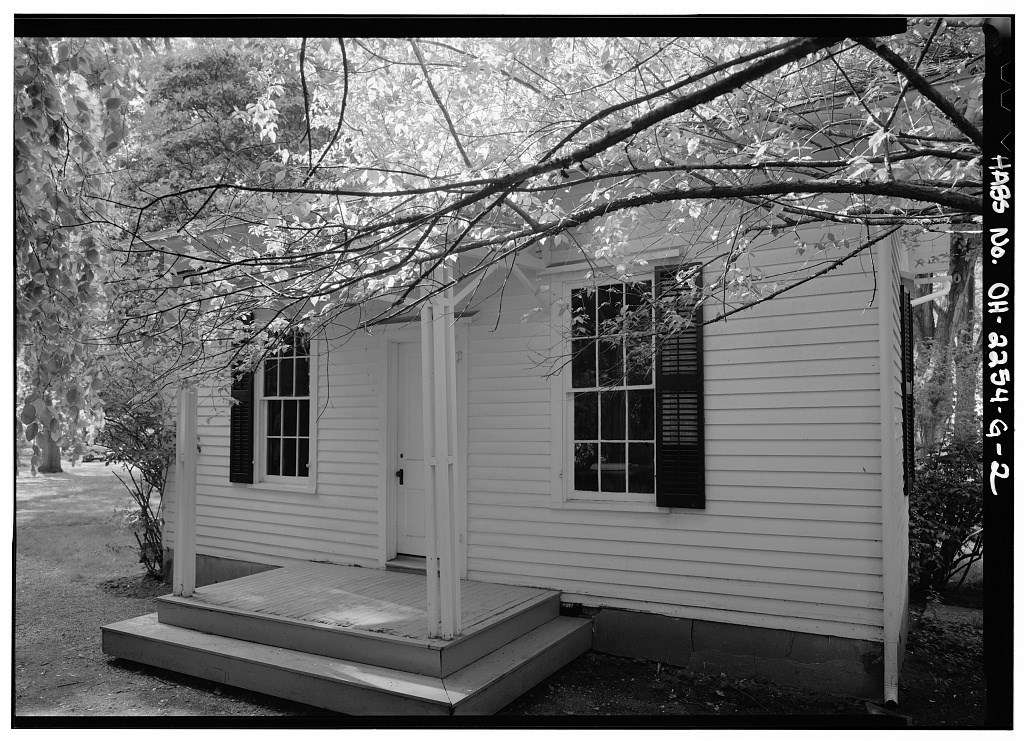 A single story structure has a small porch with a roof supported by two pillars and a window with shutters in either side.