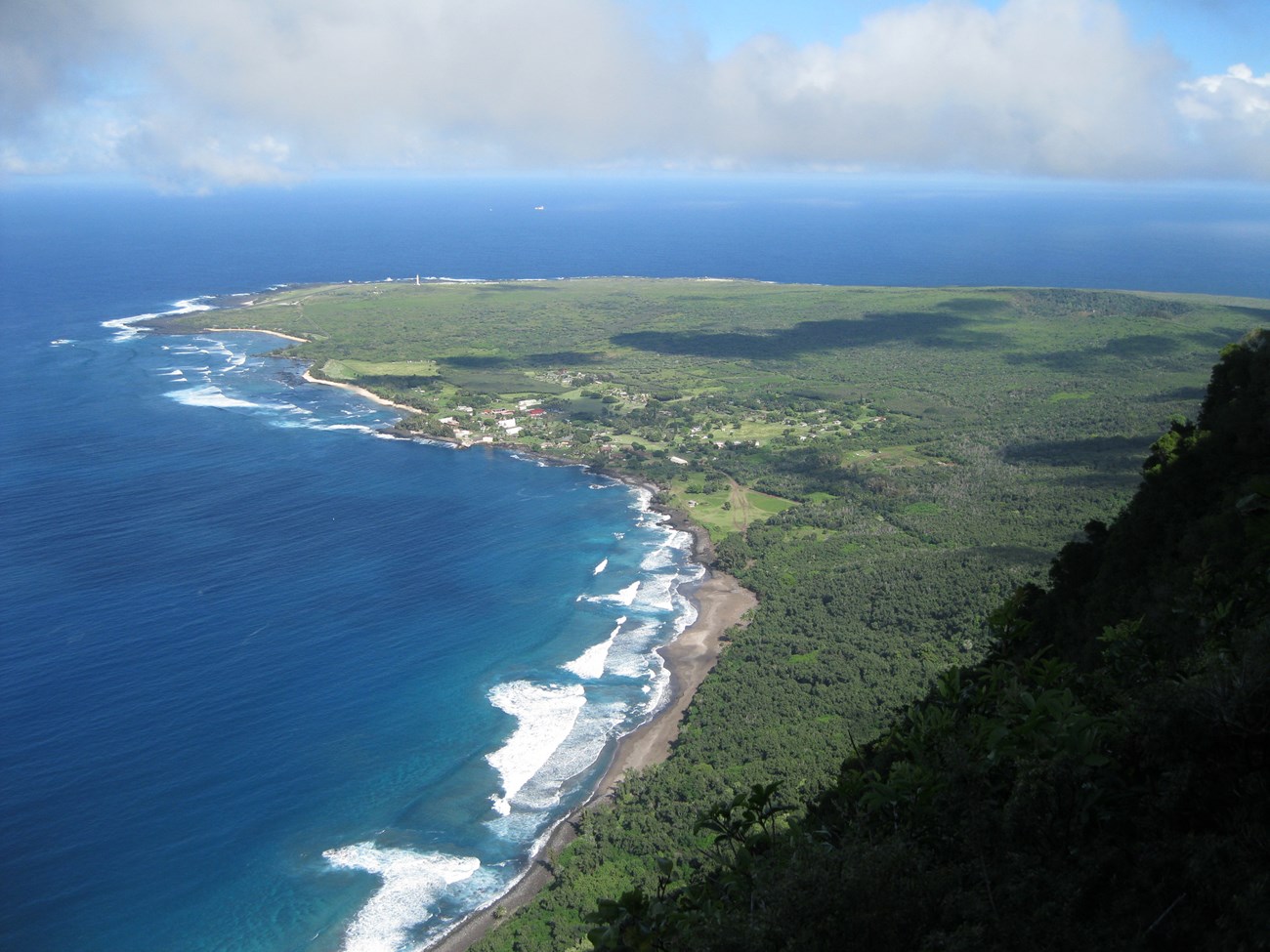 Bright blue water surrounds three sides of a green peninsula, seen from a distant cliff