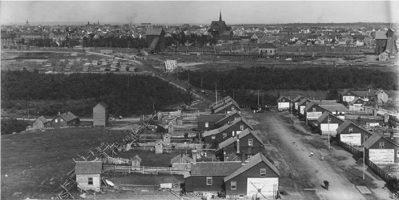 Rows of uniform houses line a street, each with a fenced yard and outbuidlings. Industrial mining structures stand in the distance, near other residential and commercial structures.