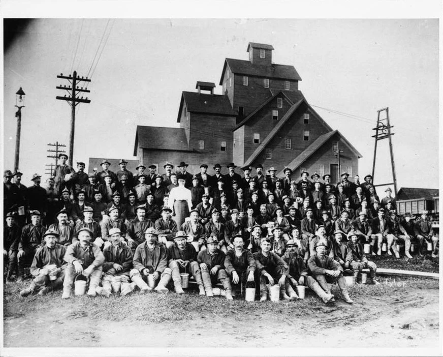 Workers at No. 2 Shaft-Rockhouse, ca. 1895-1905 A large group of workers pose in five rows, seated and standing, in front of a tall industrial shafthouse.