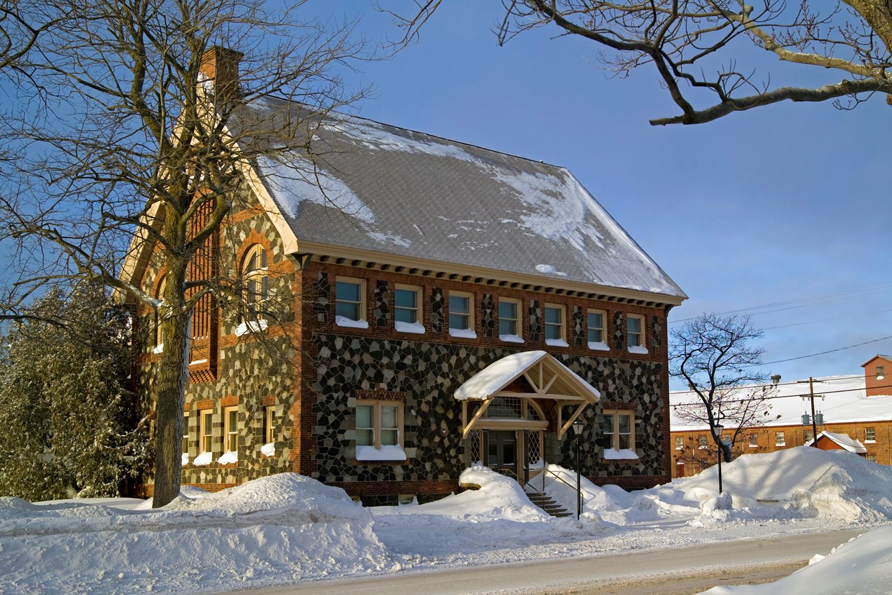 A two-and-a-half story building of mosaic, multi-toned stone from the mine is surrounded by snow and several leafless trees.