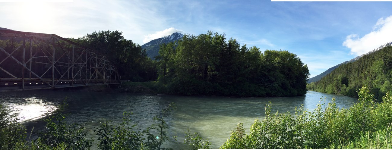 A curving river is framed by tree-covered hillsides, mountains in the background, and a trestle bridge to the left.