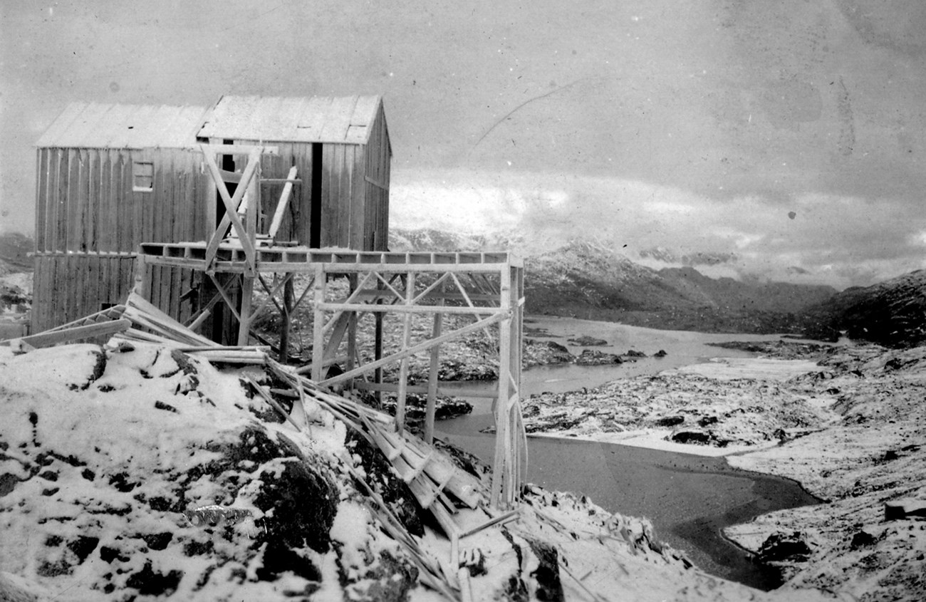 A partially collapsed wooden structure supported by beams on the edge of a snowy hillside, overlooking a lake.