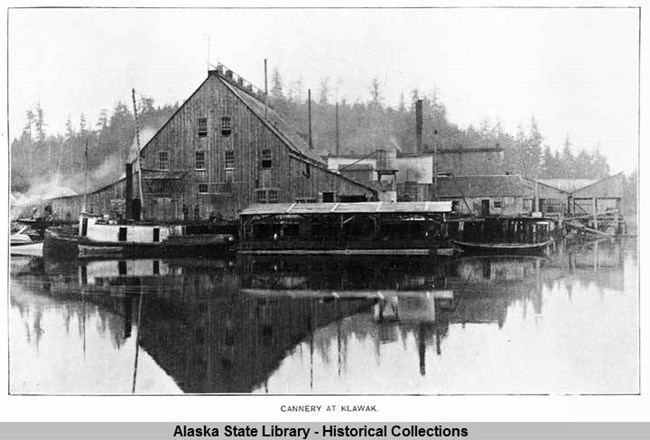 Black and white image, boats along docks and wooden warehouse-type cannery building are reflected in water, with trees and smoke in the background