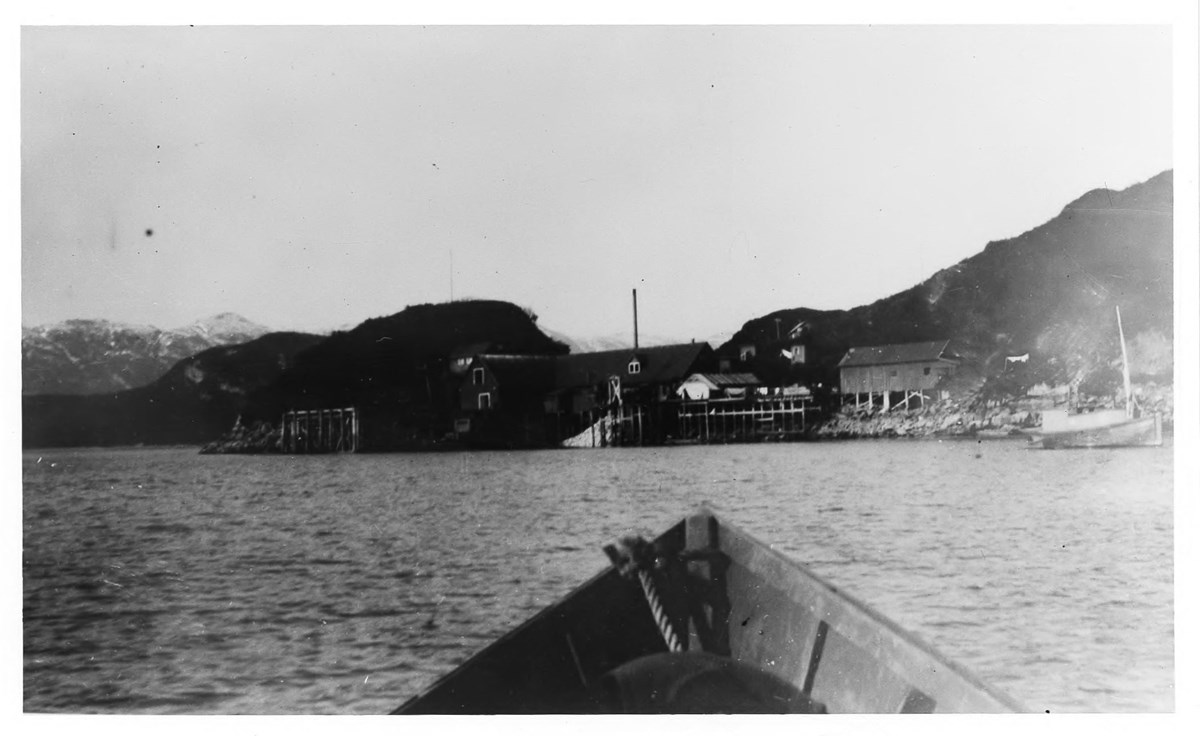 View from the bow of a boat to a collection of buildings on stilts on the coastline, black and white image