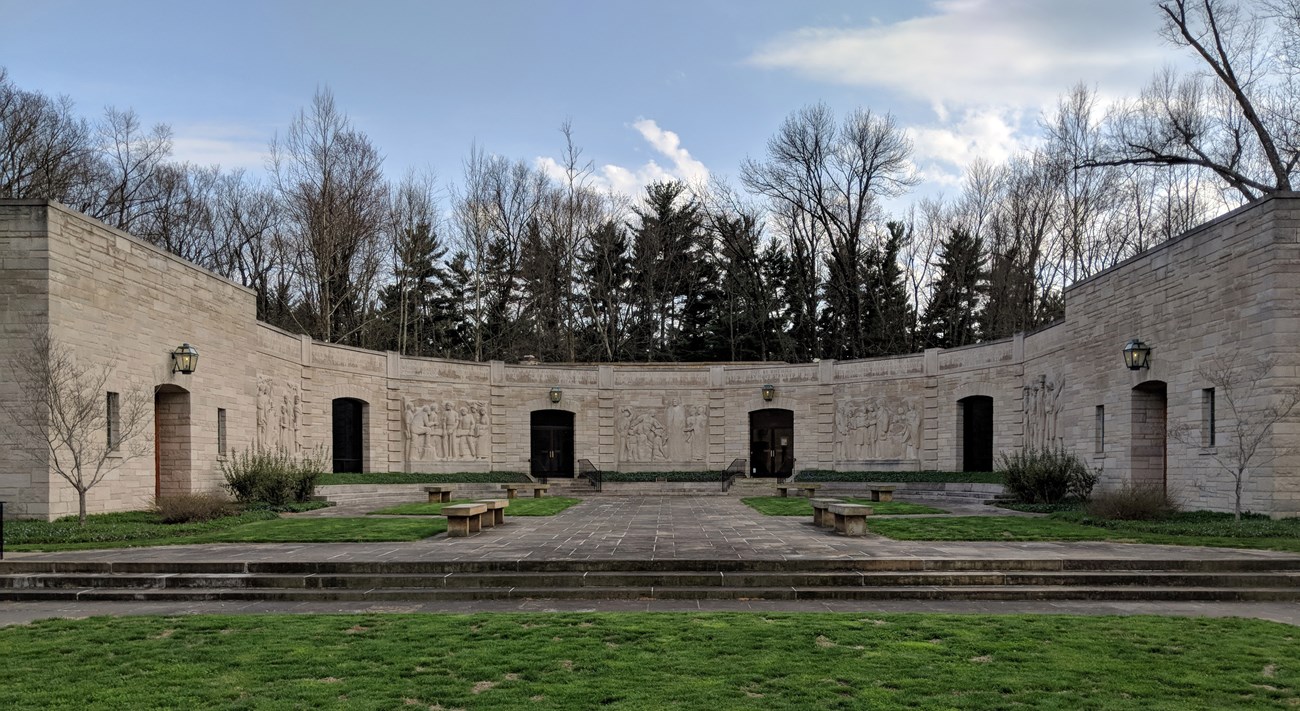 Lincoln Boyhood National Memorial Plaza Limestone block building, half circle shape with six openings spaced evenly apart. Light fixtures above four openings. Two windows on each side of left opening and right opening. Green grass in foreground leads to stairs and walkway with stone benches on
