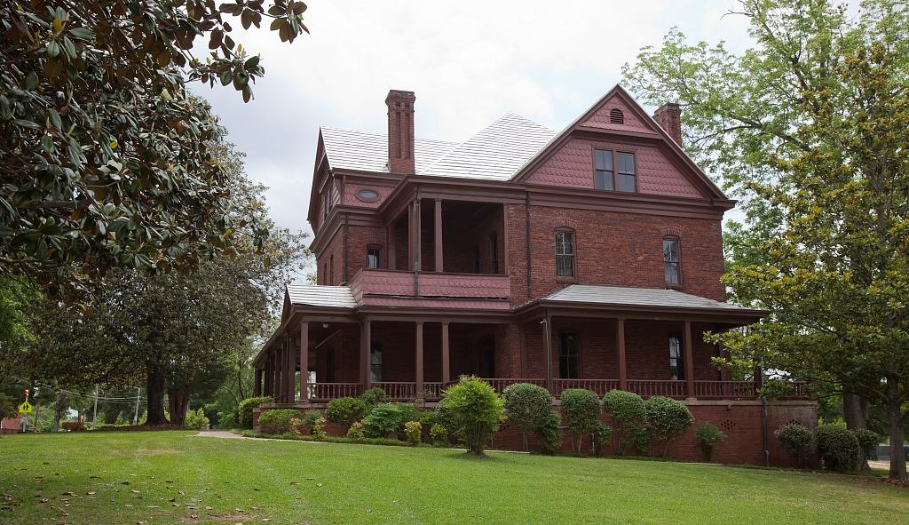 Lush lawn, mature trees, and shrubs frame the red bricks and shingles of a three-story house.