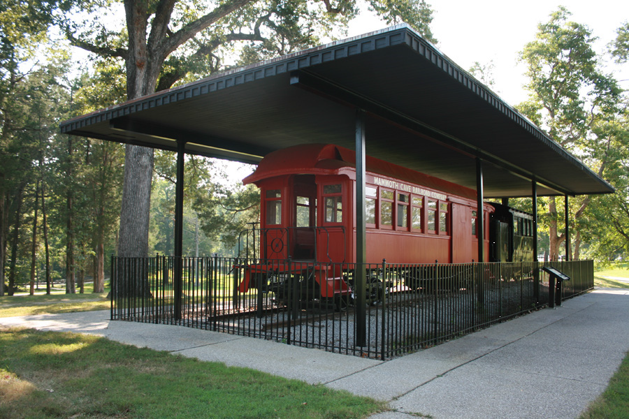 Train Exhibit at Mammoth Cave A red engine and black train coach are under a covered pavilion, surrounded by trees, sidewalk, and grass