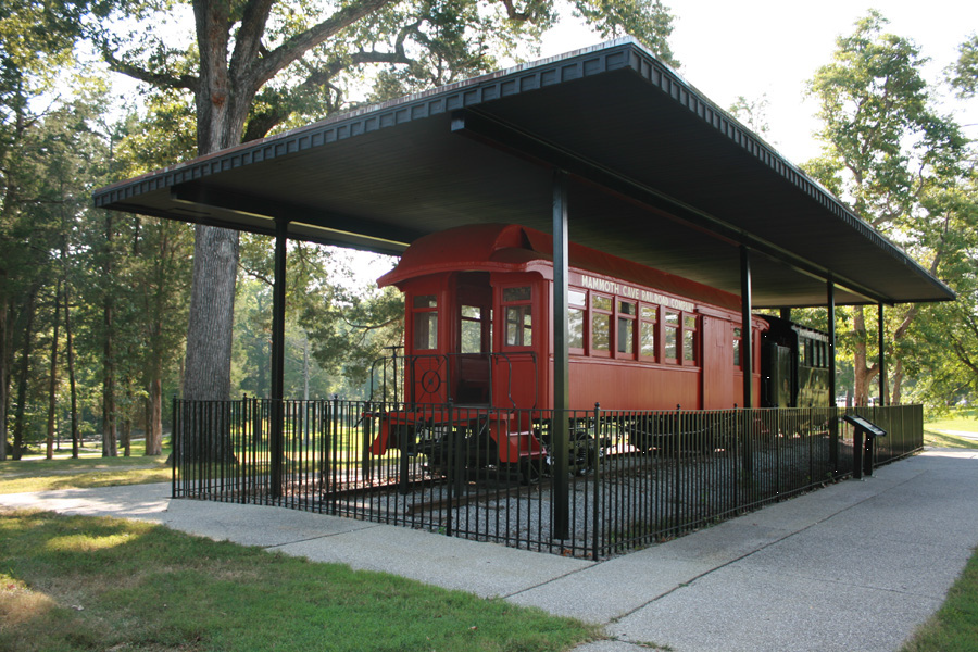 A red engine and black train coach are under a covered pavilion, surrounded by trees, sidewalk, and grass