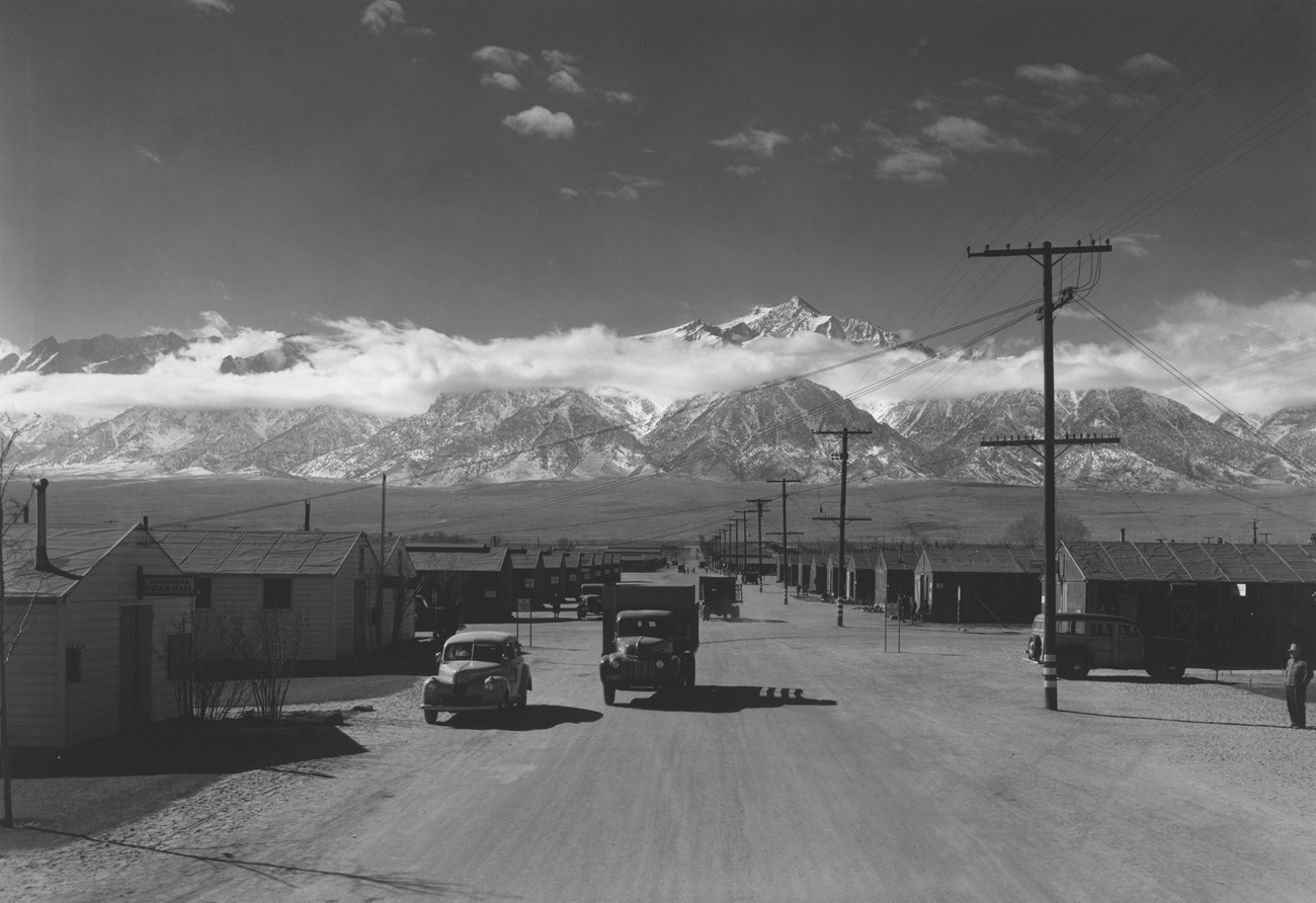 MANZ Ansel Adams 1945 Trucks drive down a wide, dusty road between rows of barracks, with mountains in the background