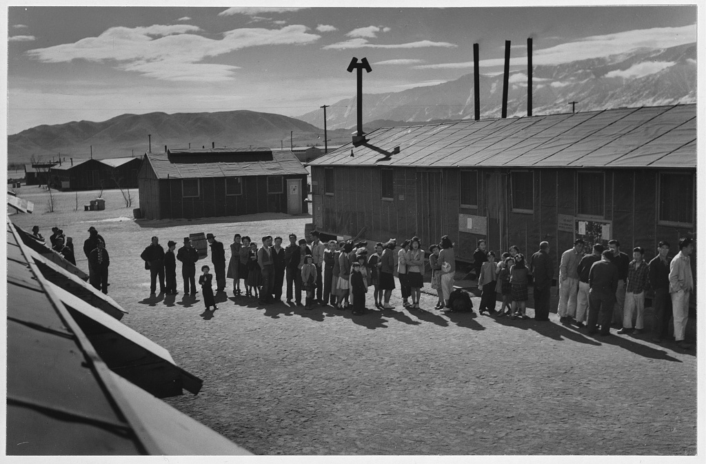 People of varying ages stand in a line in a dirt area outside a building at midday