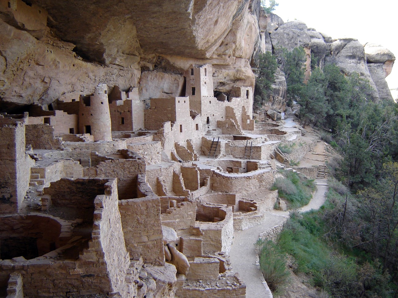 A collection of sand-colored buildings and walls with stairs and ladders is built under an overhanding cliff