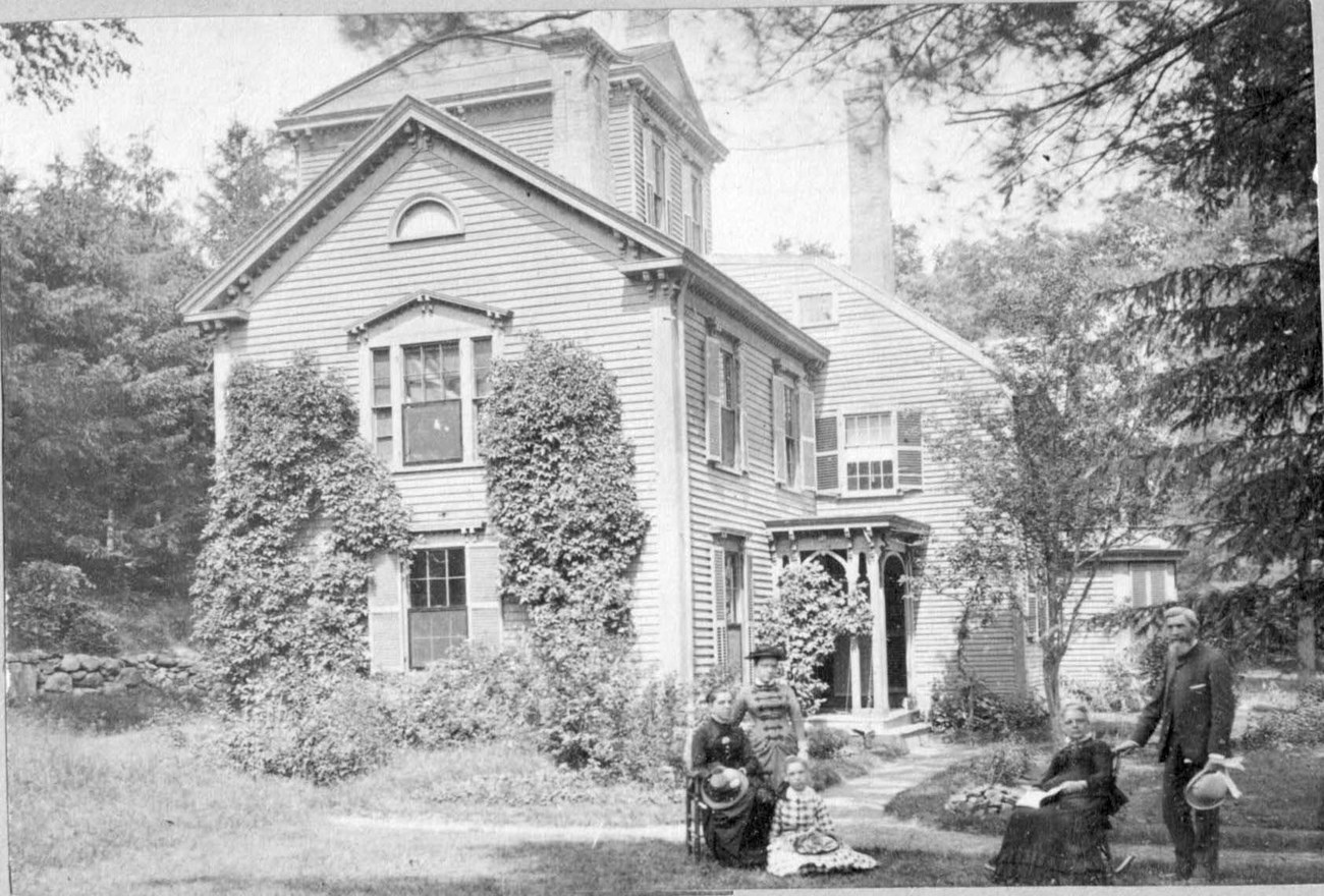 Historic photo of The Wayside from early 1880s. Five people, some seated and some standing, in a yard beside a house framed by vines and several trees in the 1880s