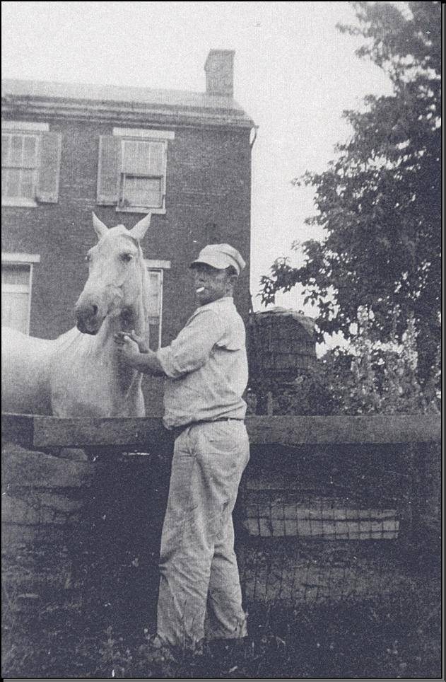 A man in a hat stands beside a horse, in front of a two-story brick building
