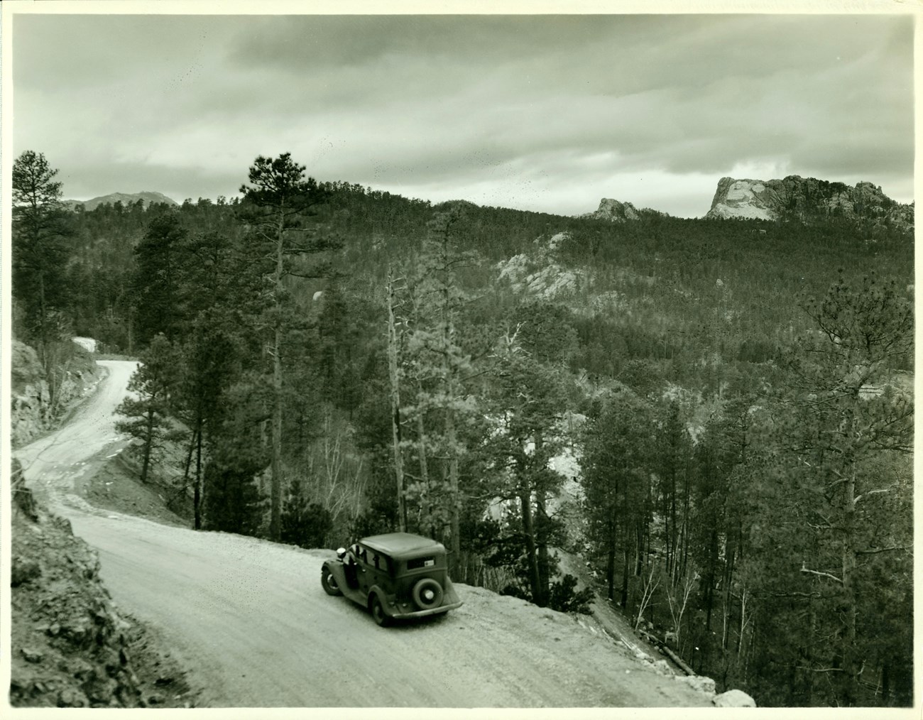 Historic photo of a car on a winding, unpaved road through a tree-covered landscape, with a rock feature rising in the distance.