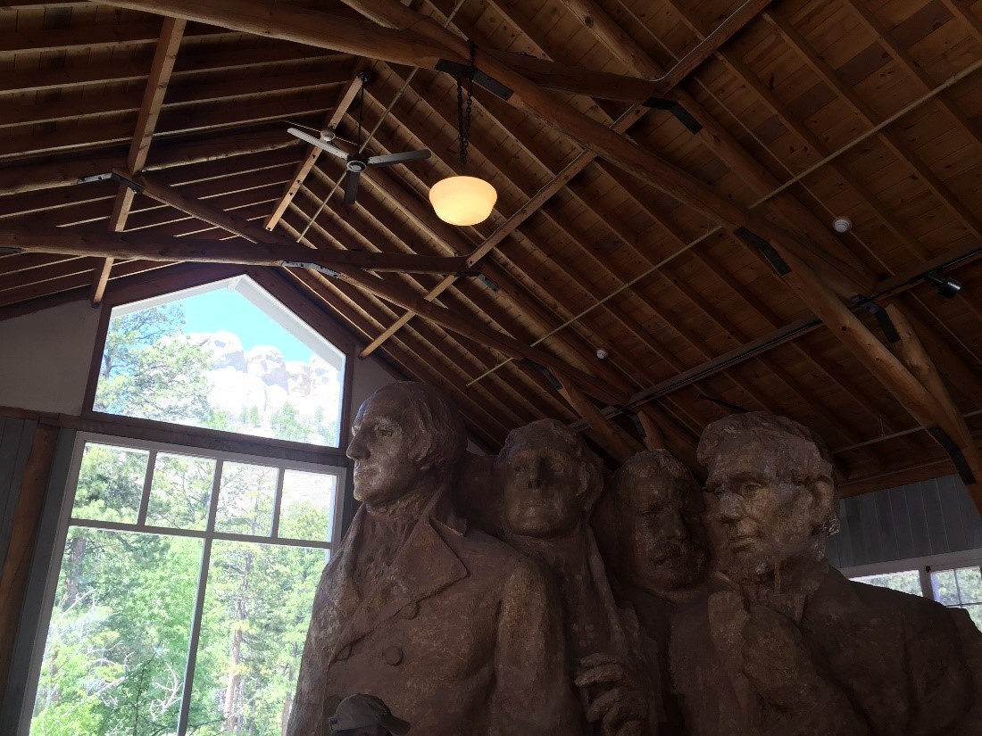 The carved faces of Mount Rushmore are visible through an opening in a wooden structure, where a model of the sculpture stands in the foreground.